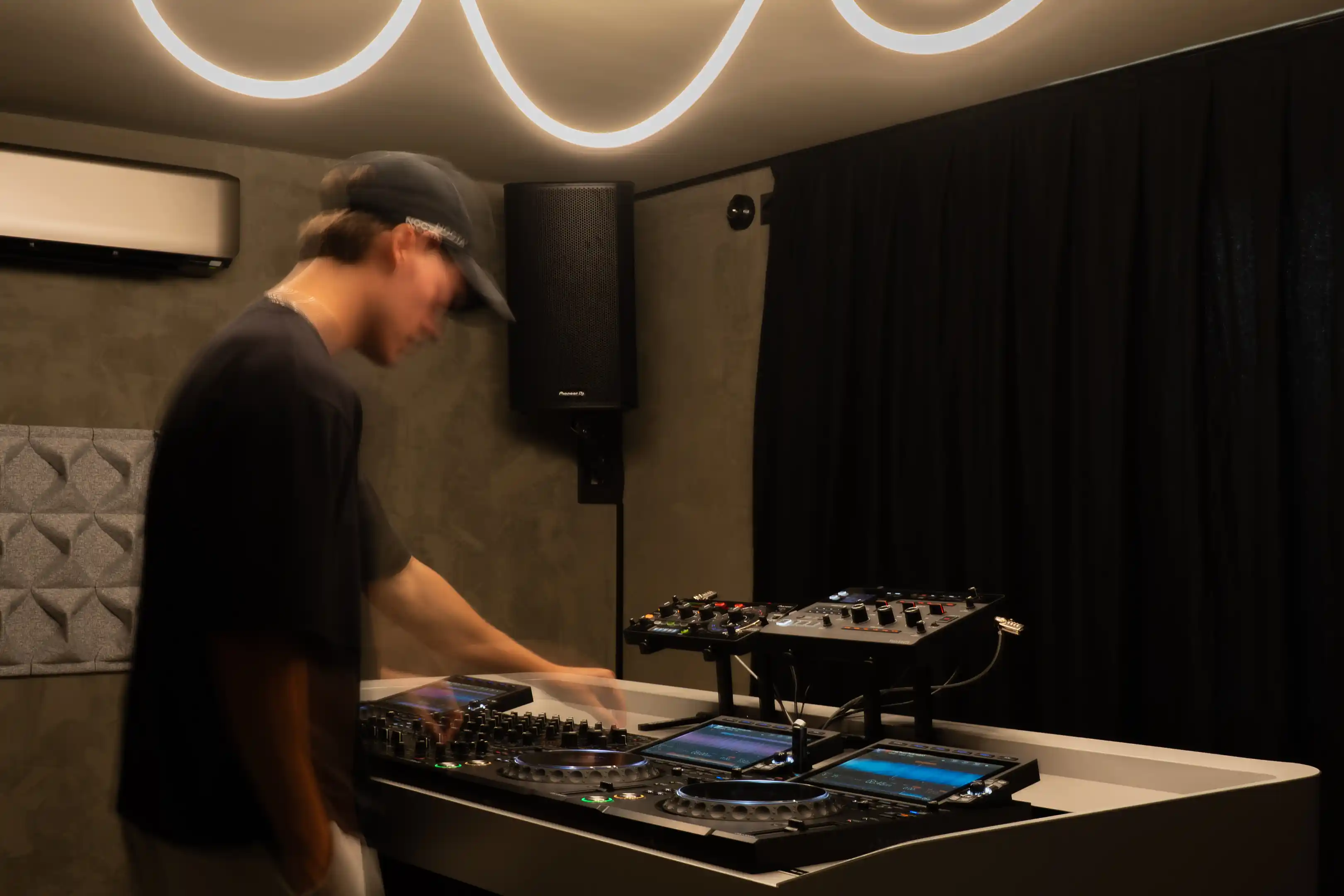 Een DJ aan het werk achter de booth, met neonlicht aan het plafond en een speaker aan de muur